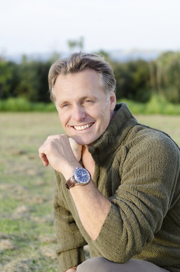 Handsome mature blond man in his forties smiling as he wears a green sweater and a brown wrist watch.