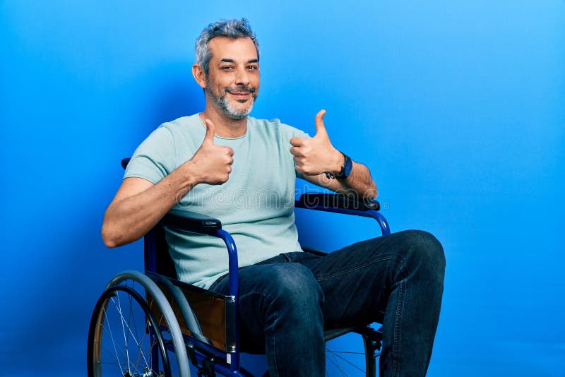 Handsome middle age man with grey hair sitting on wheelchair success sign doing positive gesture with hand, thumbs up smiling and