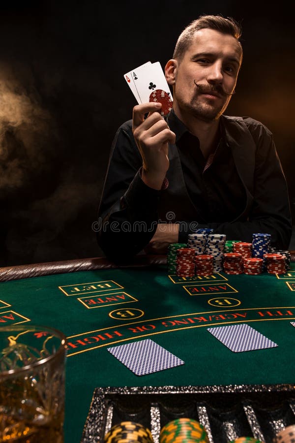 Handsome poker player with two aces in his hands and chips sitting at poker table in a dark room full of cigarette smoke stock photos Handsome poker player with two aces in his hands and chips sitting at poker table in a dark room full of cigarette smoke stock photos