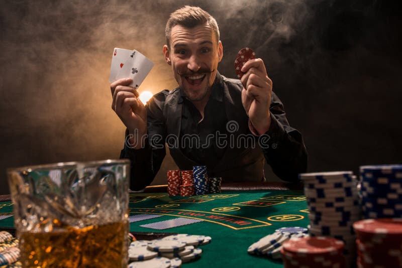 Handsome poker player with two aces in his hands and chips sitting at poker table in a dark room full of cigarette smoke royalty free stock image Handsome poker player with two aces in his hands and chips sitting at poker table in a dark room full of cigarette smoke royalty free stock image