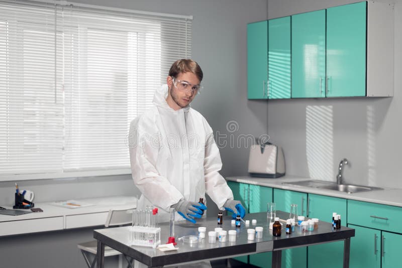 Handsome scientist wearing a white robe and safety glasses, working on something and looking at the camera stock image