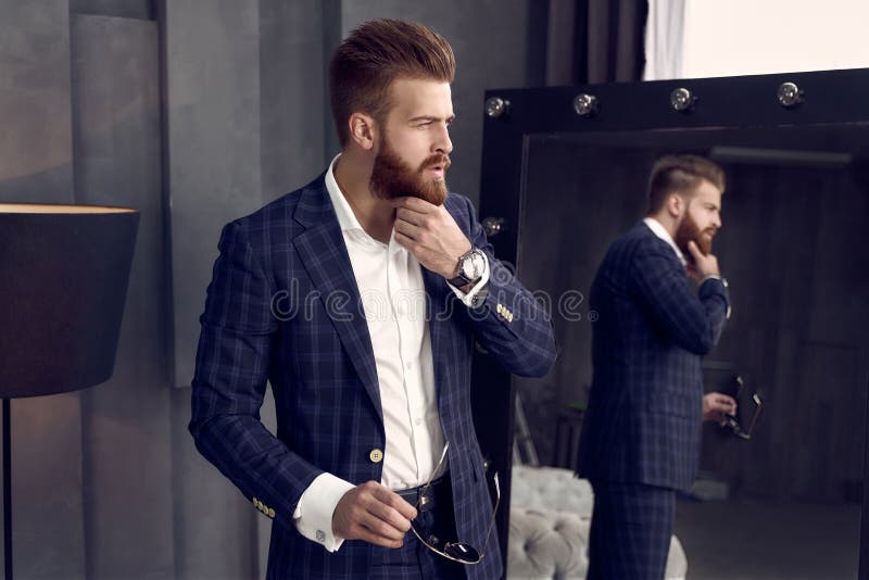 Handsome stylish man in blue suit in a cage at home standing on sofa near mirror. Holds his chin