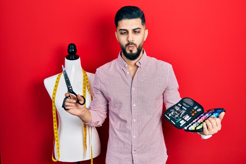 Handsome tailor man with beard holding sew kit and scissors making fish face with mouth and squinting eyes, crazy and comical royalty free stock image