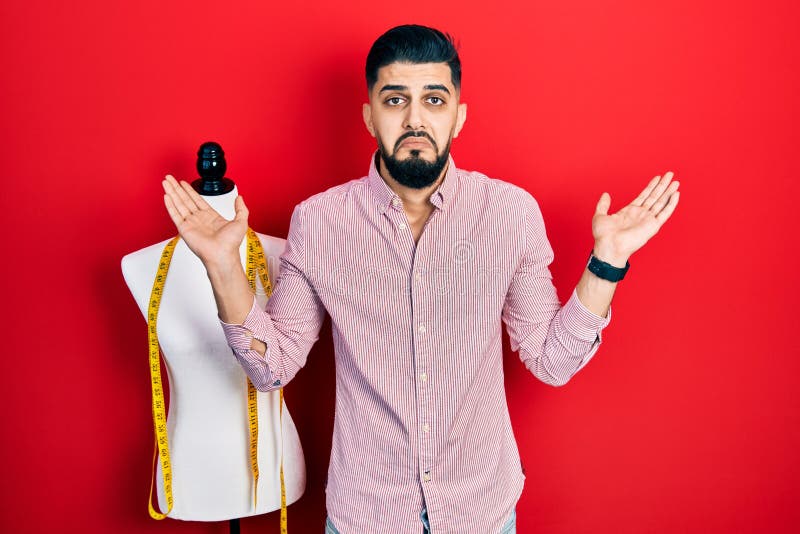 Handsome tailor man with beard standing by manikin clueless and confused with open arms, no idea and doubtful face royalty free stock images