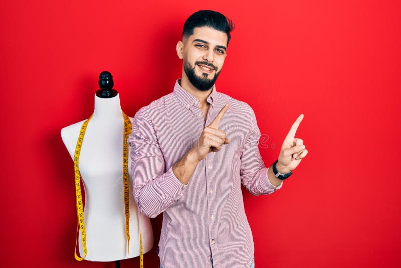 Handsome tailor man with beard standing by manikin smiling and looking at the camera pointing with two hands and fingers to the royalty free stock images
