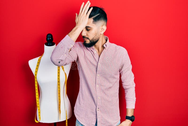 Handsome tailor man with beard standing by manikin surprised with hand on head for mistake, remember error royalty free stock photo