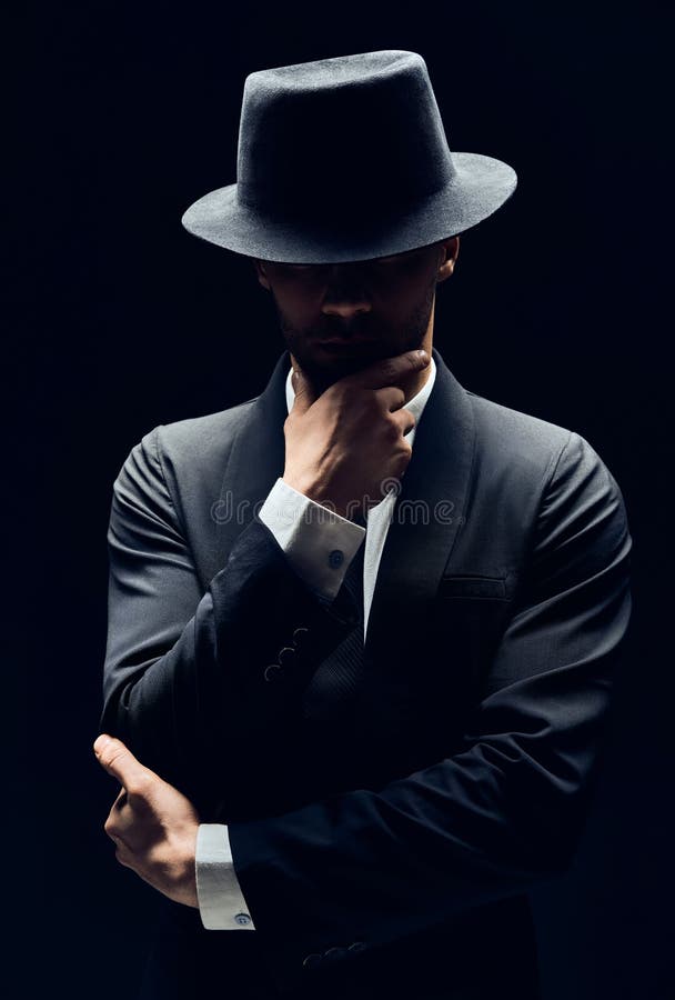 Handsome thoughtful man in black suit and hat touching chin on dark background