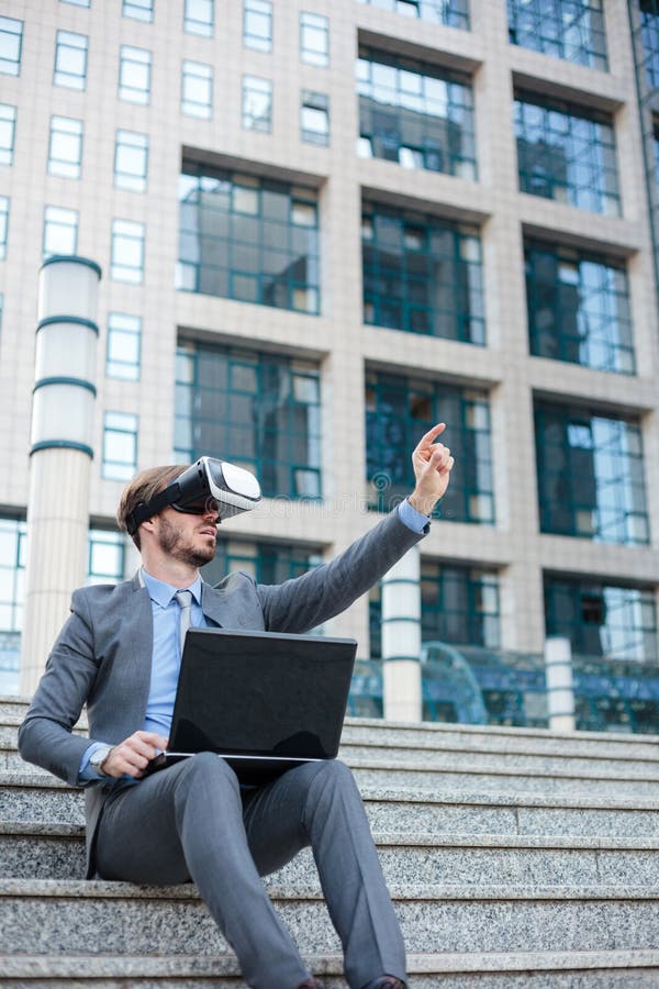 Handsome young businessman using virtual reality simulator and making hand gestures, working on a laptop in front of an office bui stock photography Handsome young businessman using virtual reality simulator and making hand gestures, working on a laptop in front of an office bui stock photography