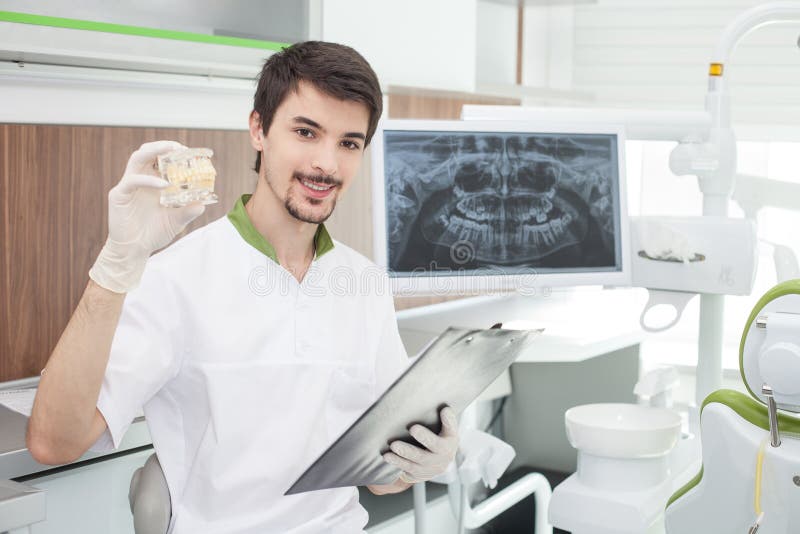 Attractive bearded dental doctor is sitting in his office. He is holding a folder of documents and smiling. The man is showing dentures and looking at camera happily. Attractive bearded dental doctor is sitting in his office. He is holding a folder of documents and smiling. The man is showing dentures and looking at camera happily