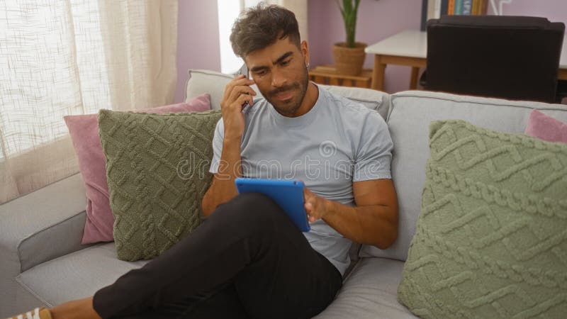 Handsome young hispanic man talking on the phone while using a tablet in a cozy living room royalty free stock photo