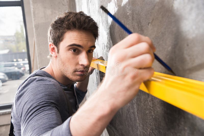 Handsome young laborer with pencil measuring wall with level tool, renovation concept