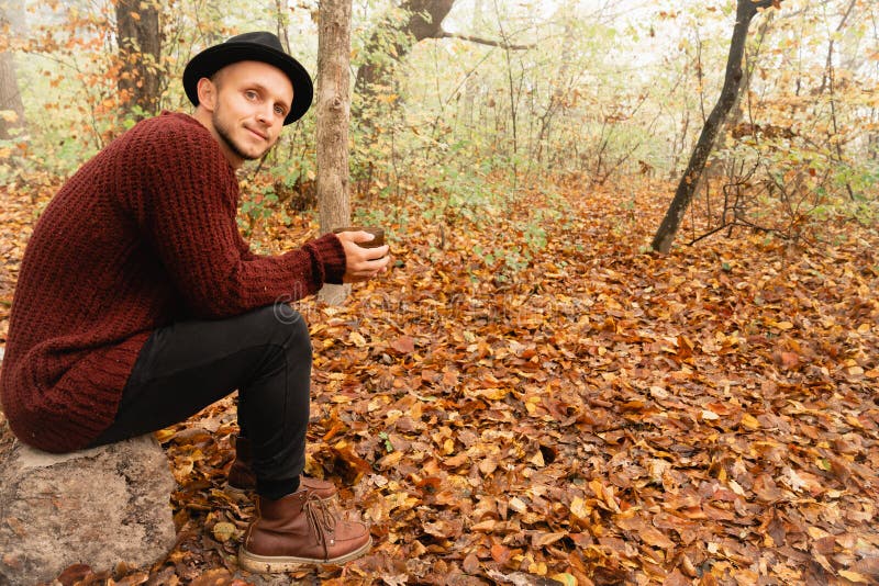 Handsome young man in black hat and red sweater sitting on the stump in the forest and enjoying morning with coffe stock photo Handsome young man in black hat and red sweater sitting on the stump in the forest and enjoying morning with coffe stock photo
