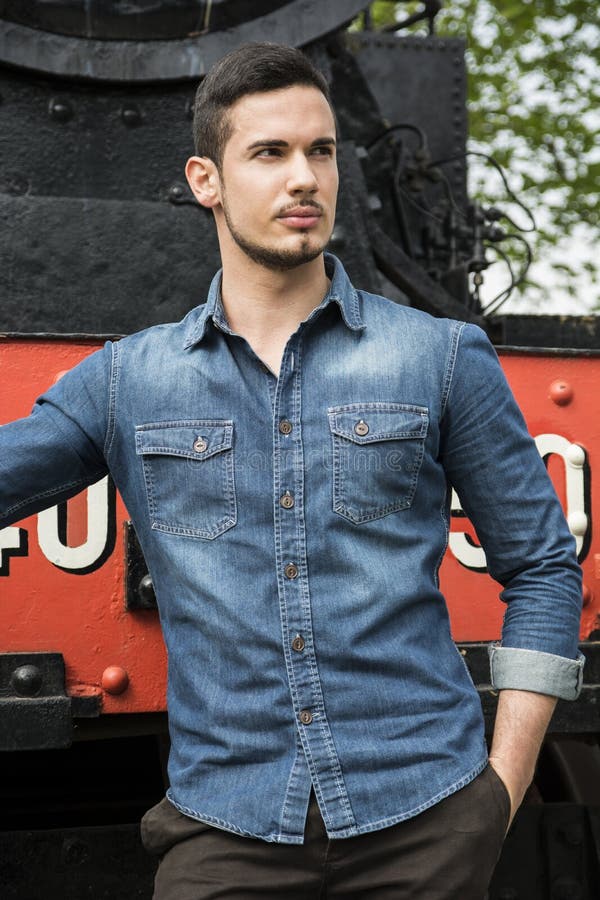 Handsome young man in denim shirt in front of old train, looking away