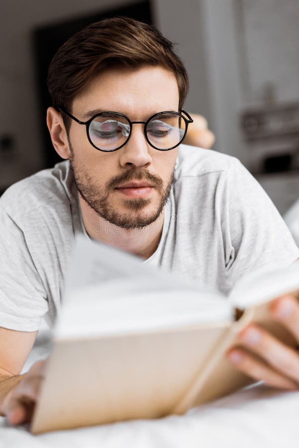 Handsome Young Man in Eyeglasses Lying in Bed and Reading Book Stock