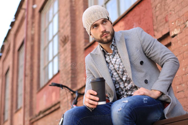 Handsome young man in grey coat and hat sitting on a bench relaxed drinking coffee and thinking near his bicycle stock photography Handsome young man in grey coat and hat sitting on a bench relaxed drinking coffee and thinking near his bicycle stock photography