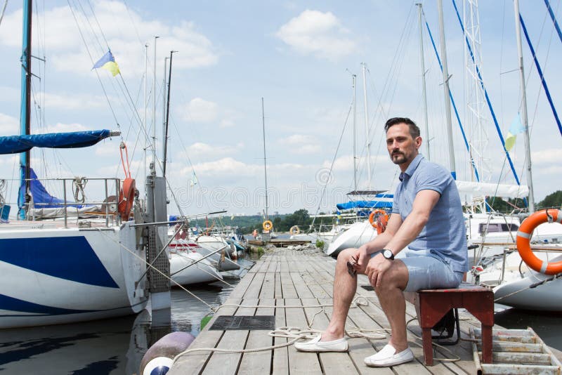 Handsome young man sitting on bench on pier between moored boats. Young businessman on vacation on pier stock photography Handsome young man sitting on bench on pier between moored boats. Young businessman on vacation on pier stock photography