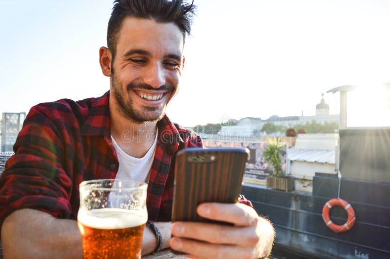 Handsome young man smiling looking at the phone and drinking a beer in a bar outside stock image Handsome young man smiling looking at the phone and drinking a beer in a bar outside stock image