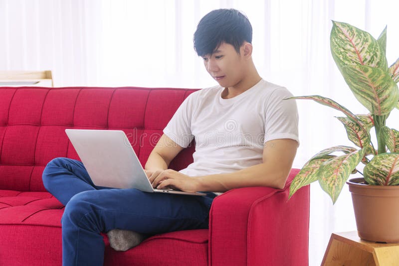 Man using laptop for working online activities while sitting on red couch at home. Freelance guy typing notebook stock images