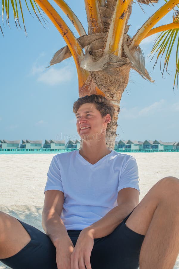 Handsome young man wearing white t-shirt and black shorts relaxing under palm tree at tropical sandy beach at island luxury resort. In Maldives stock images Handsome young man wearing white t-shirt and black shorts relaxing under palm tree at tropical sandy beach at island luxury resort. In Maldives stock images
