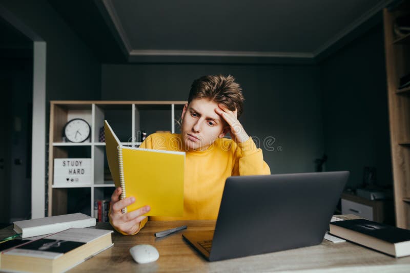 Handsome young man in yellow sweatshirt reads notebook while sitting at table at home in bedroom. The teenager is studying