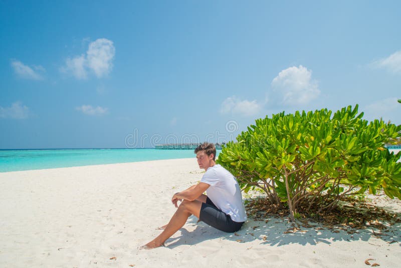 Handsome young model man in white t-shirt sitting on sandy beach at tropical island luxury resort. In Maldives stock photo Handsome young model man in white t-shirt sitting on sandy beach at tropical island luxury resort. In Maldives stock photo