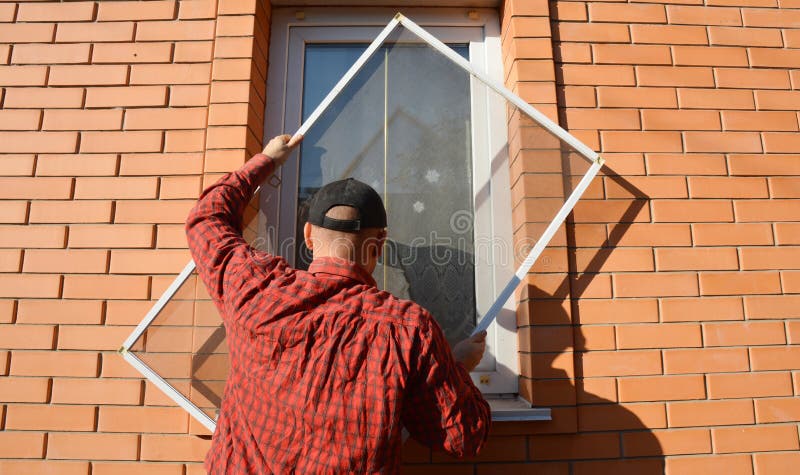 A handyman is installing anti-insect mosquito net, screen, fly and bugs protection on a plastic window of a brick house to keep stock photography