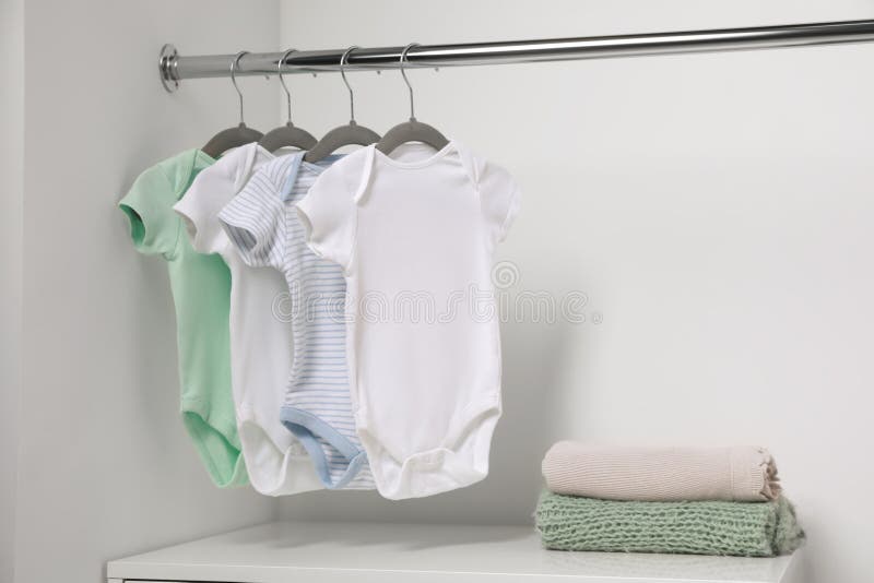 Hangers with Baby Bodysuits and Stack of Clothes Near White Wall. Space