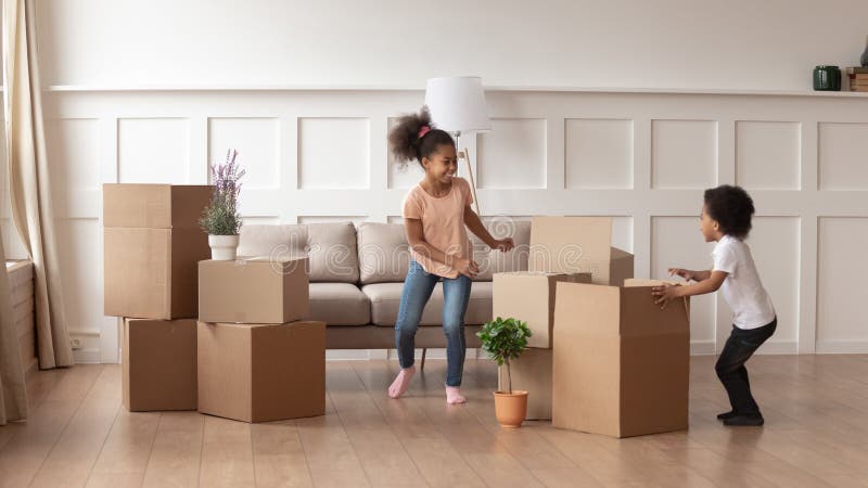 Happy active african american kids playing on moving day, two excited funny cute little mixed race children boy and girl running around cardboard boxes laughing in living room in new house apartment. Happy active african american kids playing on moving day, two excited funny cute little mixed race children boy and girl running around cardboard boxes laughing in living room in new house apartment