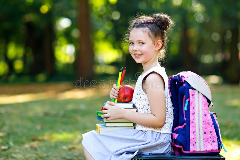 Happy adorable little kid girl reading book and holding different colorful books, apples and pencils on first day to school or nursery. Back to school concept. Healthy child of elementary class