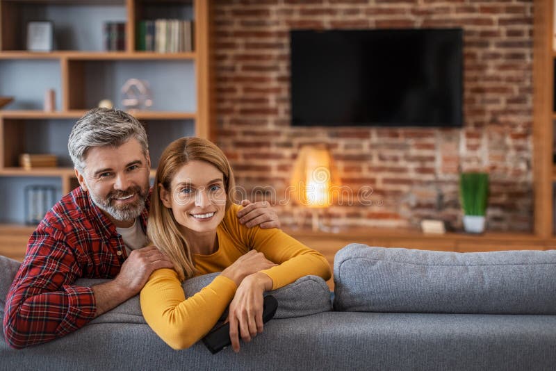 Happy adult european man and woman looking at camera, enjoy relaxing in living room interior with tv with blank screen