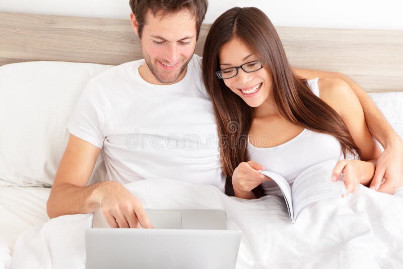 Happy affectionate young married couple sitting side by side in bed relaxing and laughing at information on the screen of their laptop. Happy young interracial couple, Asian woman, Caucasian man. Happy affectionate young married couple sitting side by side in bed relaxing and laughing at information on the screen of their laptop. Happy young interracial couple, Asian woman, Caucasian man.