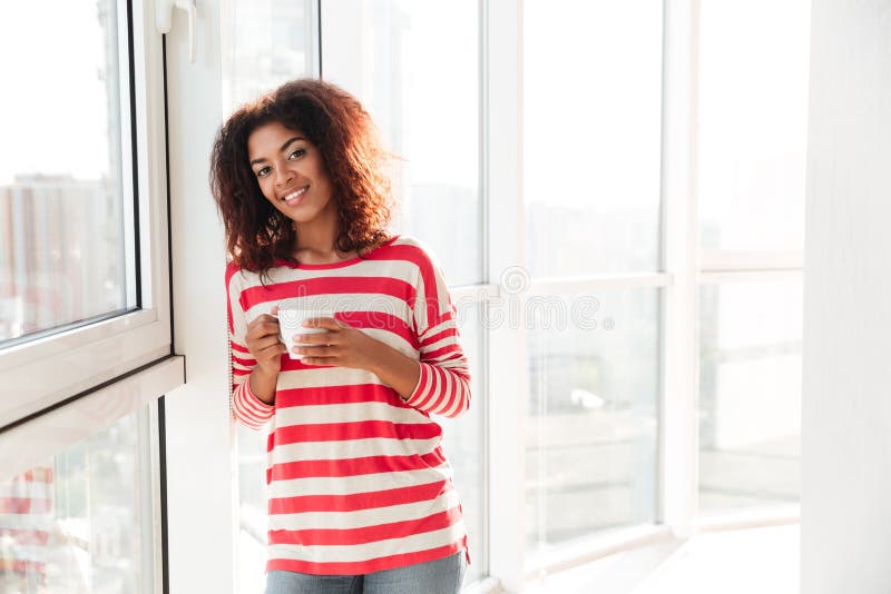 Happy african woman drinking coffee near the window