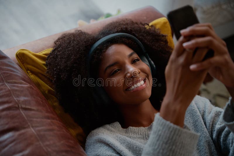 Happy african woman with headphones listening to music on smart phone lying on a couch at home royalty free stock image