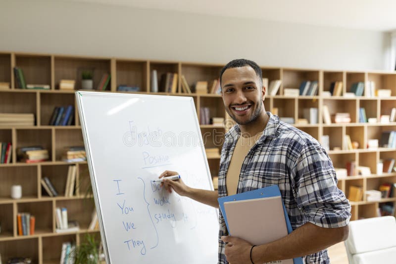 Happy arab male teacher writing rules on whiteboard, looking and smiling at camera in living room interior royalty free stock photo