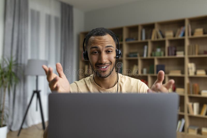 Happy arab man in headphones having video call on laptop, talking and gesturing at computer webcamera in home office royalty free stock photo