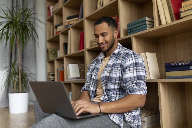 Happy arab man typing on laptop keyboard while leaning on bookshelf and studying or working from home royalty free stock images
