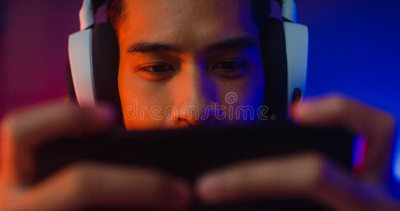 Happy asia girl gamer wear headphone competition play video game online  with smartphone colorful neon lights in living room at home. Esport  streaming game online, Home quarantine activity concept. Stock Photo