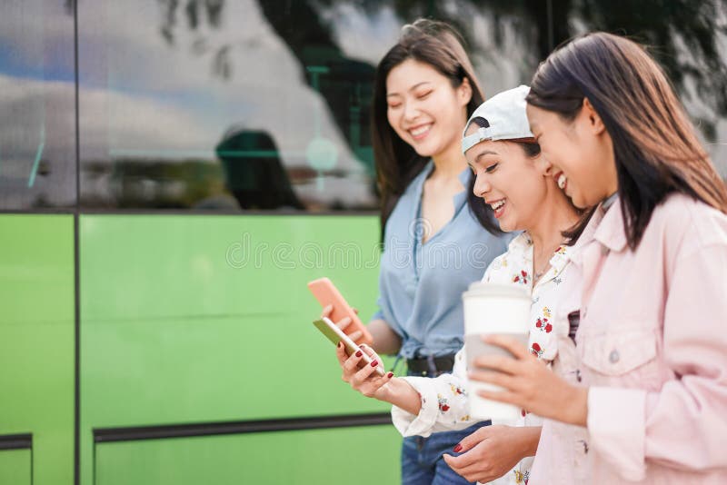 Happy asian friends using smartphones at bus station - Young students people having fun with phones app after school - Friendship stock photo