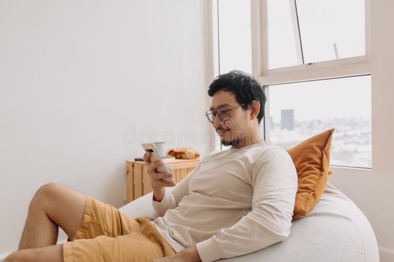 Happy asian man in long sleeve sit relaxed on bean bag using smartphone royalty free stock photos