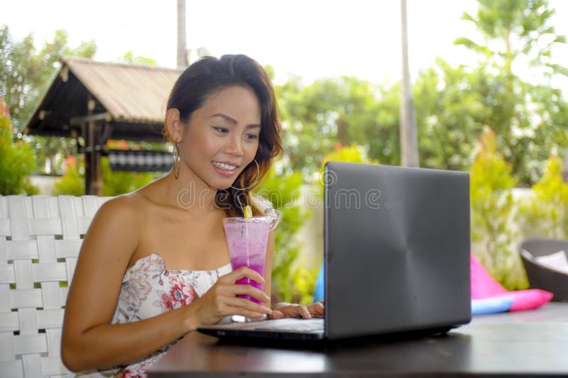 Happy Asian woman in elegant and dress sitting outdoors at pool resort coffee shop having healthy fruit juice working on lapt