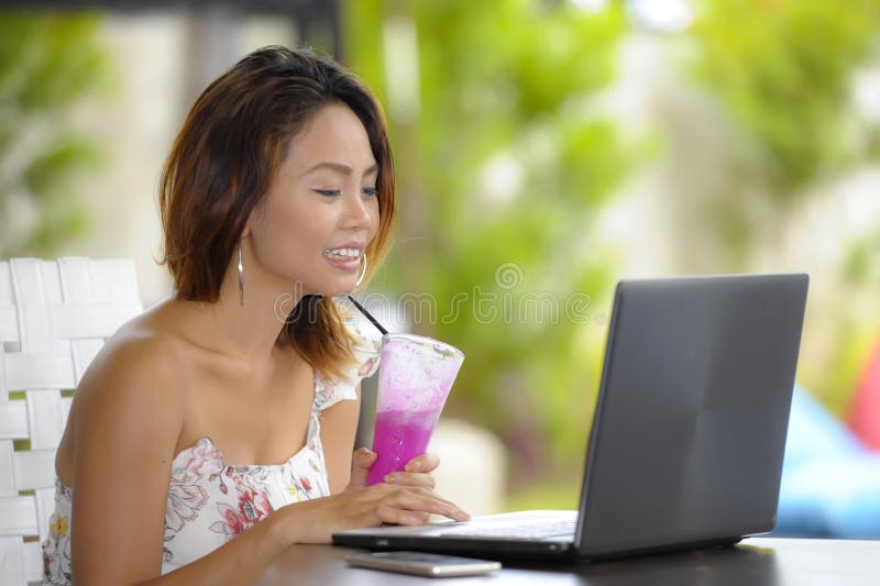 Happy Asian woman in elegant and dress sitting outdoors at pool resort coffee shop having healthy fruit juice working on lapt