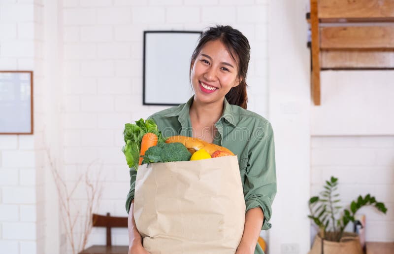 Happy asian woman  holding vegetable bag walking into kitchen after shopping at grocery store at home.vegan lifestyle concept