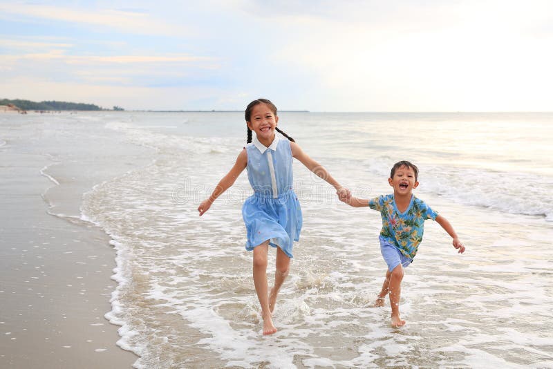 Happy Asian young sister and little brother having fun running together on tropical sand beach at sunrise. Happy family boy and