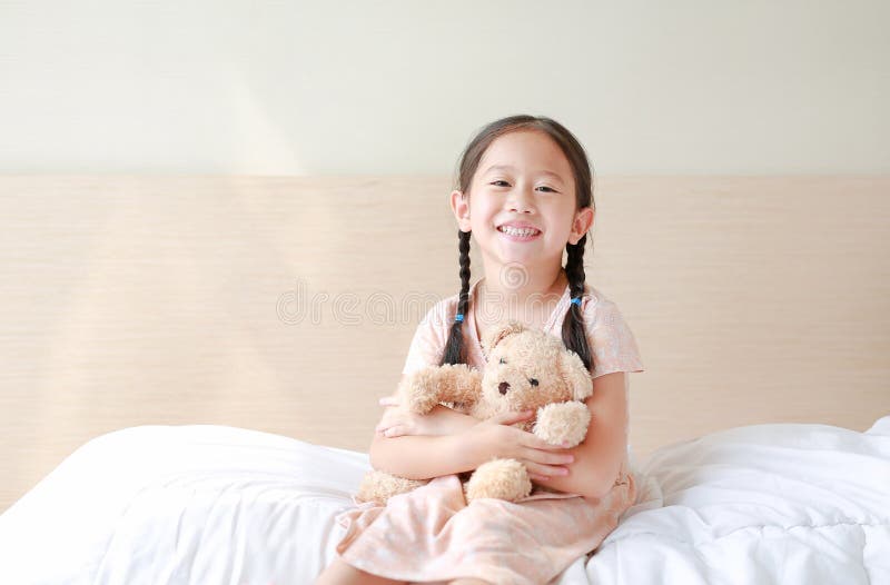 Happy asiatisches kleines Mädchen, das Teddybären umarmt und Kamera ansieht, während es zu Hause auf dem Bett sitzt stockfoto Happy asiatisches kleines Mädchen, das Teddybären umarmt und Kamera ansieht, während es zu Hause auf dem Bett sitzt stockfoto