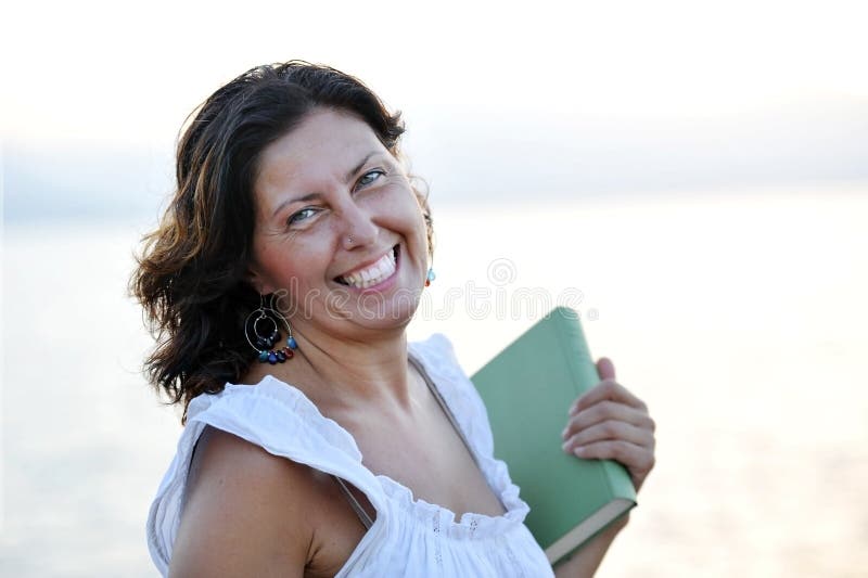 mature woman on her 40s holding a book and smiling at the beach outdoors, portrait in front of the sea enjoying a relaxing summer evening in calm. mature woman on her 40s holding a book and smiling at the beach outdoors, portrait in front of the sea enjoying a relaxing summer evening in calm