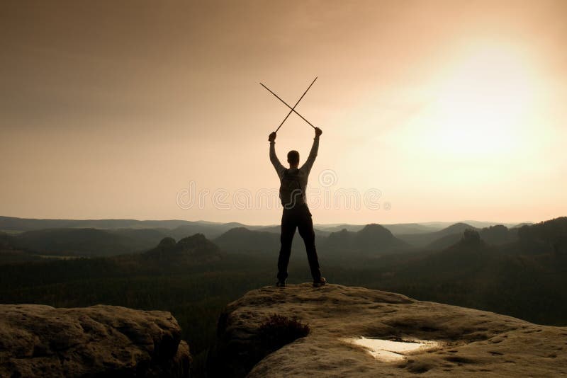 Happy backpacker with x crossed poles in the air, open misty mountain valley bellow cliff. Silhouette of tourist