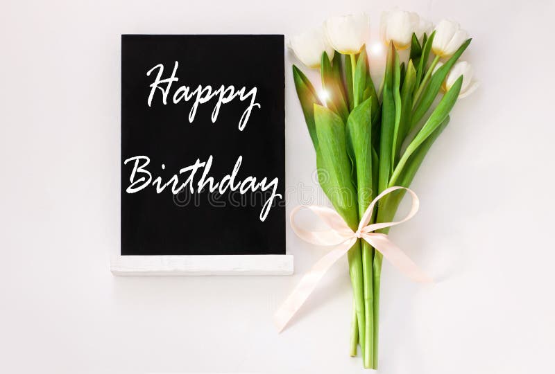 Happy Birthday card message sign on black chalkboard with tulip flowers on white background flat lay. Blackboard greeting text top view. Natural floral decoration green leaves. Celebrate party banner. Happy Birthday card message sign on black chalkboard with tulip flowers on white background flat lay. Blackboard greeting text top view. Natural floral decoration green leaves. Celebrate party banner