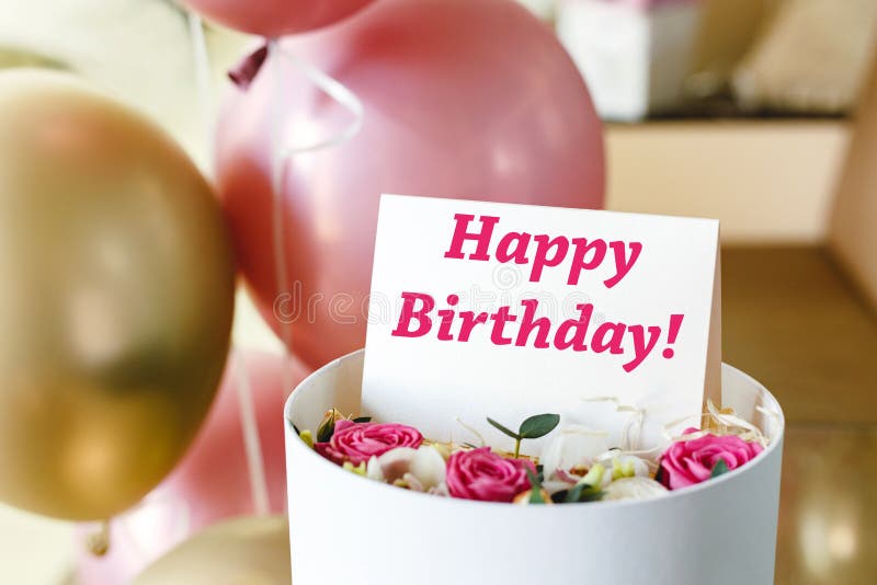 Happy Birthday text on gift card in flower box near festive pink and gold balloons. Beautiful bouquet of fresh flowers roses in box with greeting card Happy Birthday