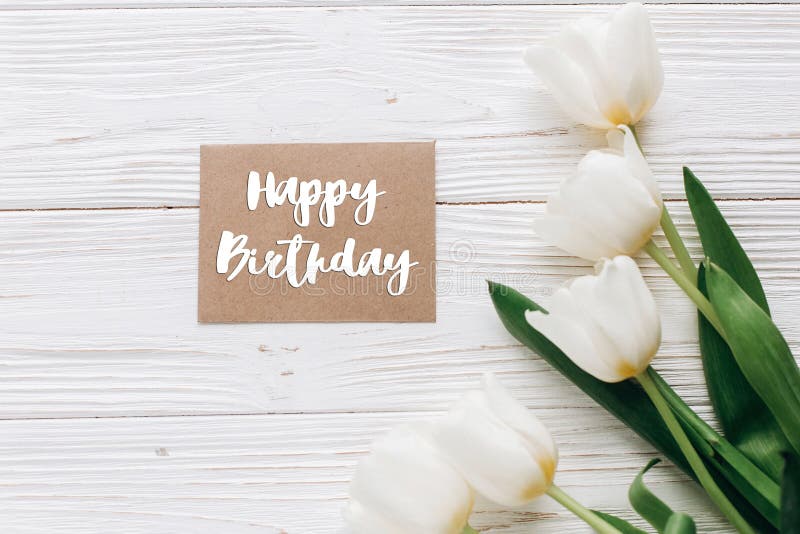 Happy birthday text sign on stylish craft greeting card and tulips on white wooden rustic background. flat lay with flowers and gift blank paper with space for text. greeting card