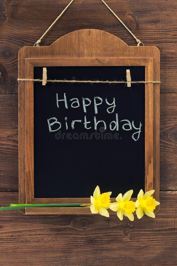 Happy Birthday written with chalk on aged blackboard with yellow narcissus flowers hanging on wooden wall